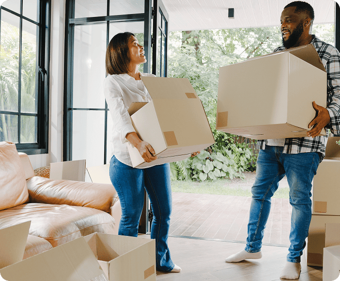 Two people stand in a living room holding cardboard boxes, surrounded by more boxes, as they move into or out of a house with large windows—an experience often praised in Brightside moving group reviews for smooth moving services.
