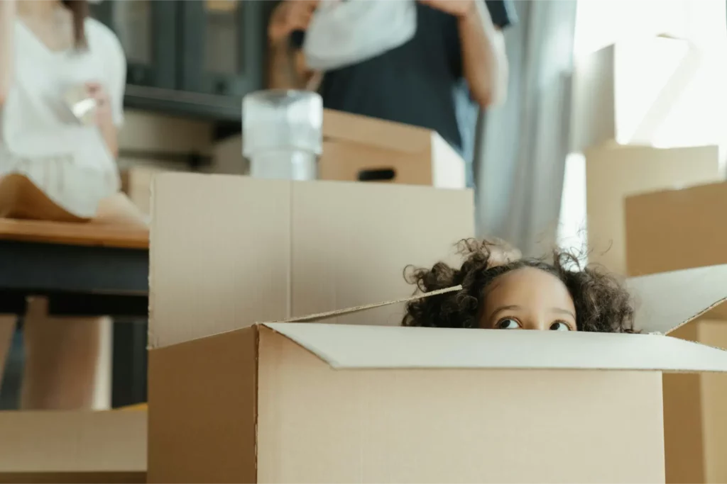 A young child peeks out from inside a cardboard box as two adults and moving boxes surround them—capturing the excitement of moving day with the help of long distance movers.