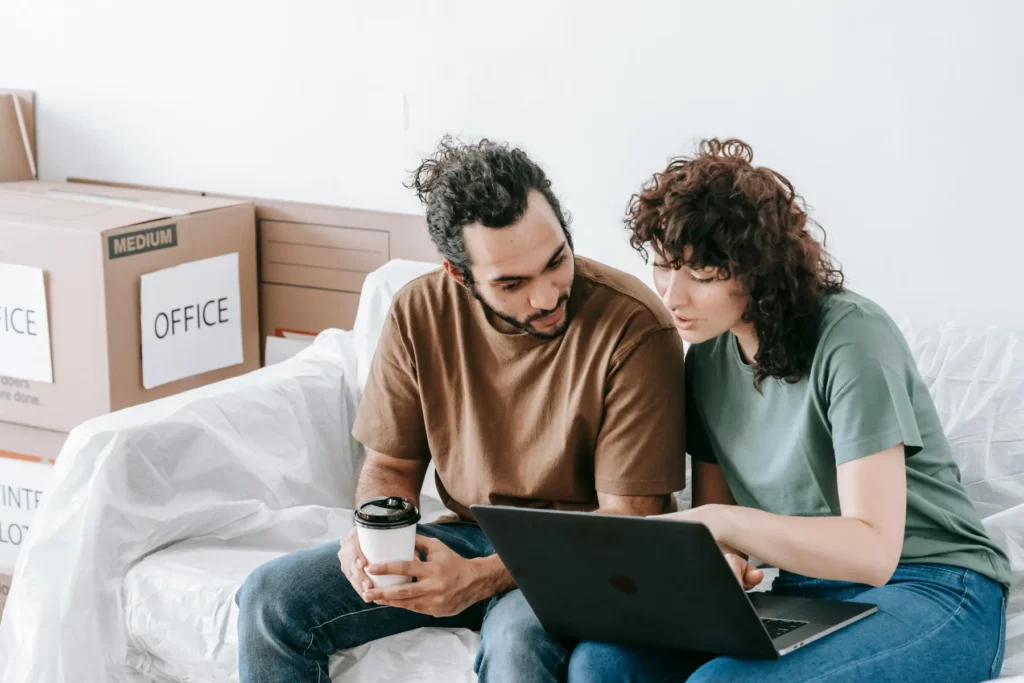 Two people sit on a couch surrounded by moving boxes labeled "OFFICE," looking at a laptop together—perhaps searching for commercial movers near me. One holds a coffee cup, planning their office relocation.