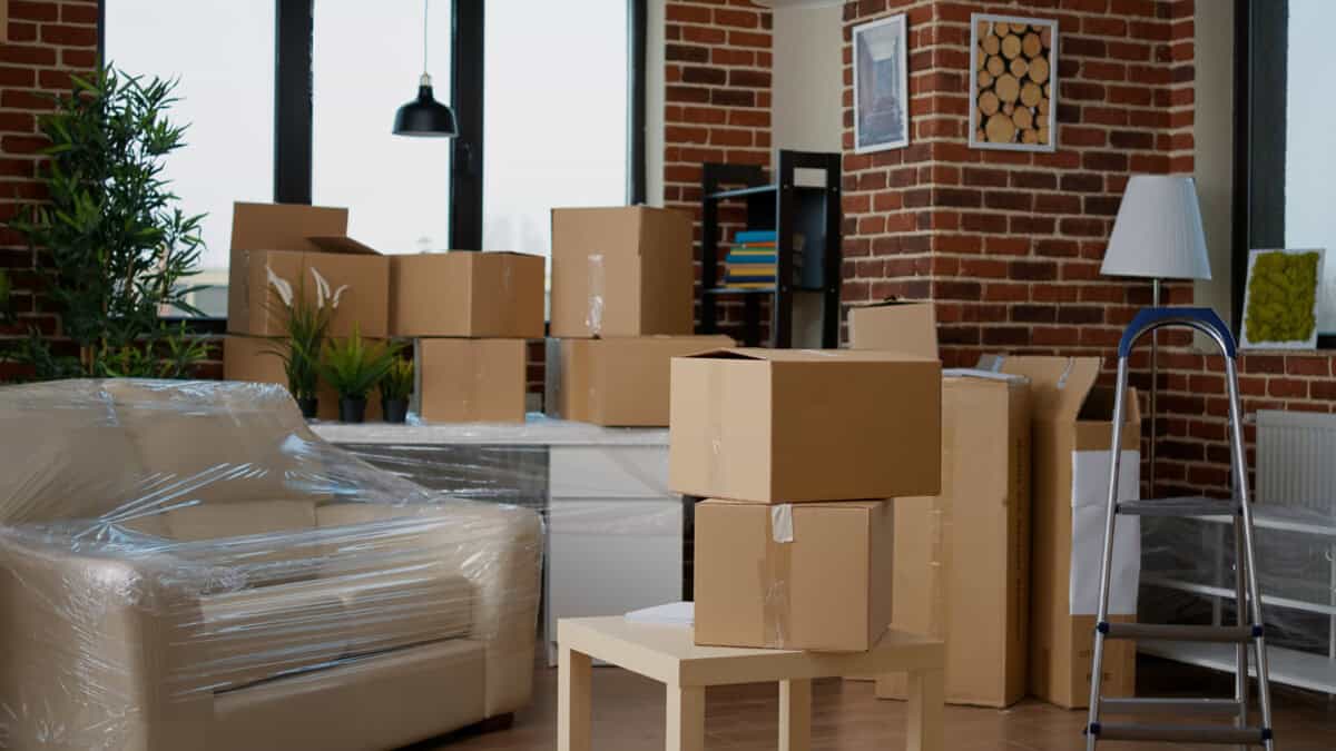 A living room with a plastic-covered sofa and several cardboard boxes stacked on the floor and furniture, suggesting a move or recent arrival—perhaps even Trevor Beekman’s new beginning.
