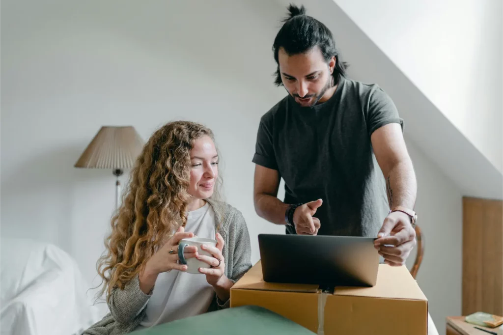 A woman holding a mug sits next to a man who is using a laptop placed on a cardboard box in a bright room, possibly reviewing a moving service estimate together.