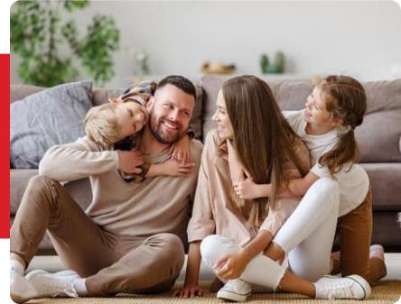 A man and woman sit on the floor with two children, all smiling and embracing, in a living room with a couch and plants in the background.