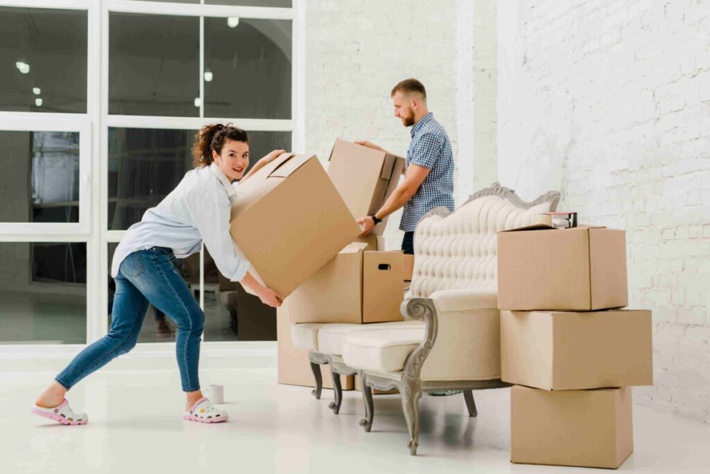 Two people are moving cardboard boxes in a bright room; one person is lifting a box while the other places a box on a sofa surrounded by more boxes.