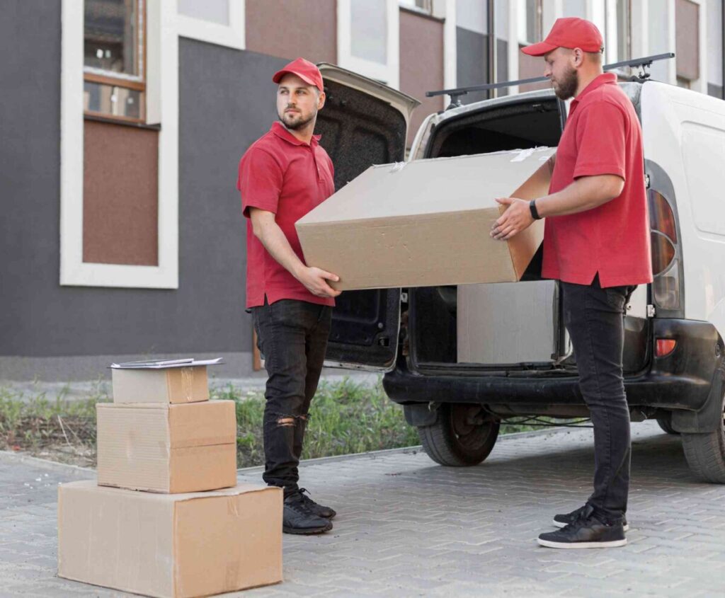 Florida Moving Services: Two delivery workers in red uniforms unload a large cardboard box from the back of a white van, with more boxes stacked nearby on the pavement.