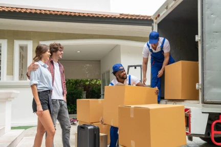 Two movers unload cardboard boxes from a truck while a couple stands nearby in front of a house, observing the process.