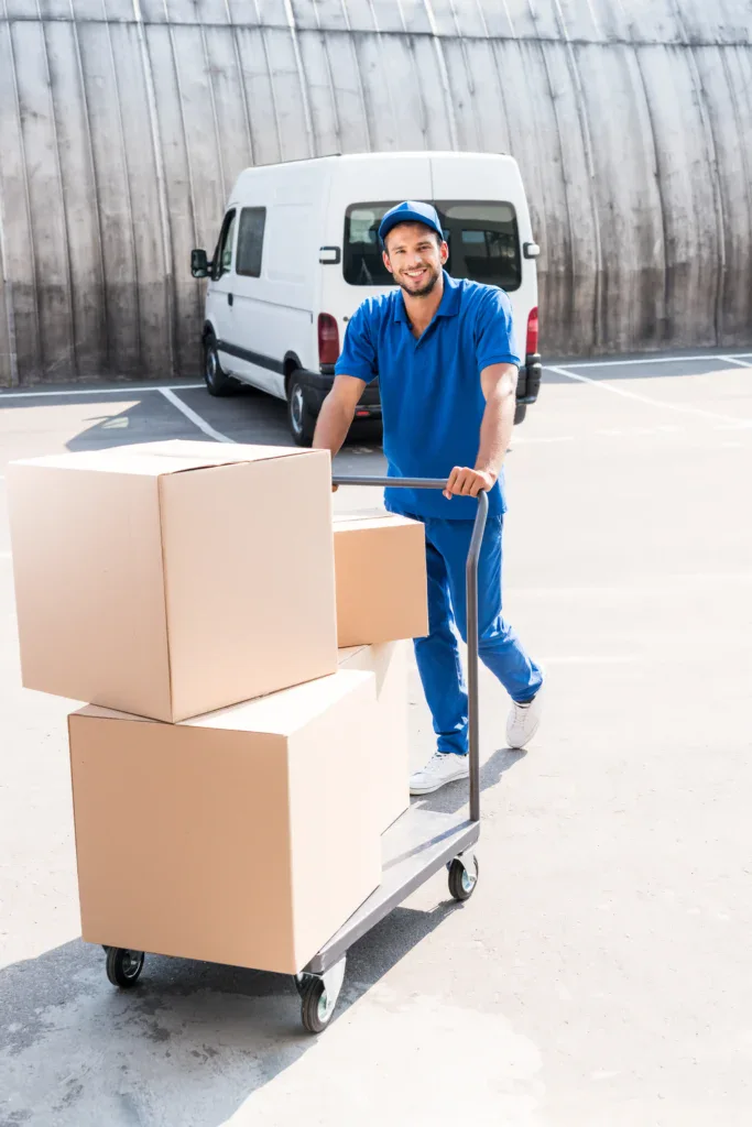 A delivery worker representing one of Brightside Moving Group's national moving companies pushes a cart with three large cardboard boxes in a parking lot.