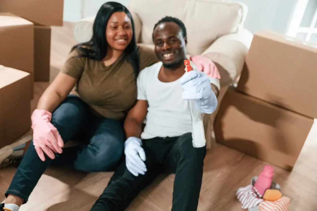 A man and woman wearing cleaning gloves sit on the floor surrounded by moving boxes and cleaning supplies, smiling and holding a spray bottle—ready to tackle their new home after using affordable movers in Florida for a stress-free, money-saving move.